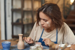 Young creative woman with paintbrush painting self-made jug or pot